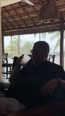 A medium indoor shot shows a man in sunglasses drinking from a frosted glass while seated in what appears to be a beach-side cabana. The man is wearing a dark t-shirt, light-colored shorts, and a wristwatch. He is positioned in the foreground, facing slightly to the right of the frame. He raises his drink with his right hand and pauses momentarily before lowering it again. The background features a thatched roof with a ceiling fan and a woven lampshade. Outside the cabana, palm trees and a beach can be seen, with rain heavily obscuring the view. The scene is somewhat dimly lit, and the man is largely in silhouette. Chairs and tables can be seen in the blurred background.