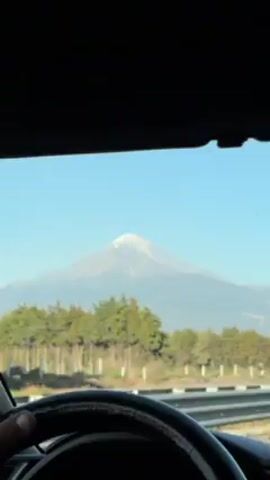 The video shows the view through the windshield of a car driving on a highway. The steering wheel is visible in the foreground. Outside the car, there is a row of trees along the highway. In the distance, there is a large mountain with a snow-capped peak. The sky is clear and blue.