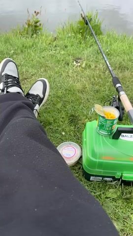 A person is sitting by a pond, fishing. The camera shows a first-person perspective of the person’s lap, sneakers, and fishing equipment. There is a fishing rod propped up in the grass next to a green tackle box. A can of corn sits on top of the tackle box, and a container of bait is nearby. The pond is calm and still, reflecting the cloudy sky. Trees line the far bank of the pond, and power lines are visible in the distance. A person can be seen walking along the opposite side of the pond.