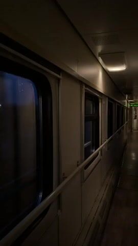 The video shows a dimly lit corridor inside a train. The walls are white, and windows line one side of the corridor, reflecting the outside light. A handrail runs along the wall beneath the windows. The floor is dark and indistinct. Overhead, a few rectangular light fixtures illuminate the passage. A small green sign is visible further down the corridor, near the end. There are no people visible in the frame. The train appears to be in motion.