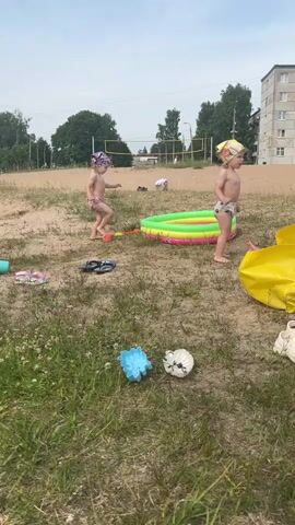 The video shows a sunny outdoor scene with two young children playing in and around a small, inflatable kiddie pool filled with water. The children are wearing swim diapers and hats. The pool is set up on a sandy area with some grass in the foreground. Scattered around the pool are children's toys and flip-flops. In the background, there is a volleyball net and a multi-story building. One child is standing in the pool and appears to be interacting with the other child, who is standing just outside the pool. A large yellow inflatable object, possibly a pool float or mattress, is visible on the right side of the frame.