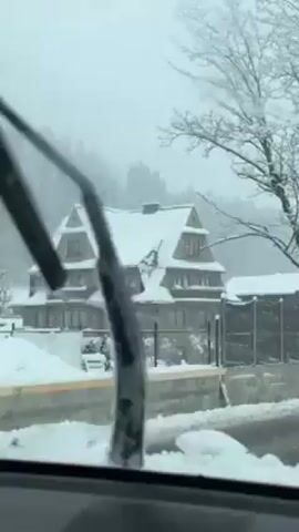 The video shows a large, multi-story house covered in snow. The house has a distinctive roof with multiple gables. Snow is falling heavily, and the surrounding trees and landscape are also covered in snow. A car windshield and windshield wipers are visible in the foreground, suggesting the video was taken from inside a moving vehicle. A gray concrete wall and a black privacy fence can be seen around the house. The overall scene depicts a winter day in a snowy, possibly mountainous, region.