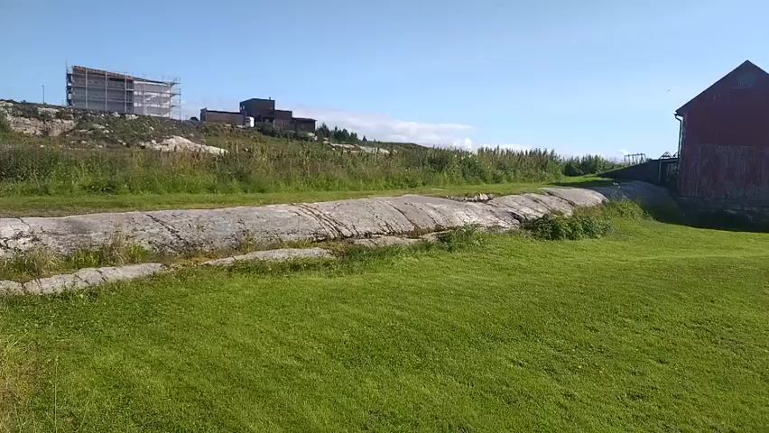 The video shows a drone flying over a grassy field towards a red building. The building appears to be a barn or a storage shed with a steep roof. In the background, there are some modern buildings under construction on a rocky hill. The sky is clear and blue with some scattered clouds. The overall scene is one of a rural landscape on a sunny day.