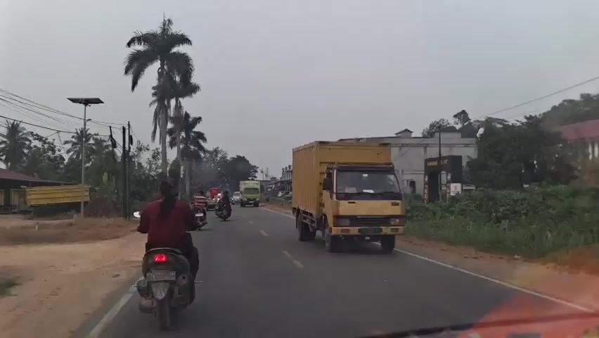 The video shows a view from a vehicle traveling on a road. A motorcycle with a rider wearing a maroon shirt is in the foreground on the left. Several other motorcycles and a green truck are on the road ahead. Buildings line the right side of the road, and palm trees are visible on the left. The sky is overcast.