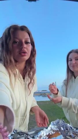 Two young women in matching cream-colored zip-up hoodies stand side-by-side, facing each other, and play rock-paper-scissors in front of the open trunk of a vehicle.  A clear blue sky is above them. In the background, a body of water, possibly a river or lake, is visible, along with a grassy area and some buildings in the distance. In front of the women, on the open trunk, is aluminum foil, and beneath it, what appears to be food. Both women are smiling and engaged in the game. One woman has curly brown hair while the other has straight brown hair.