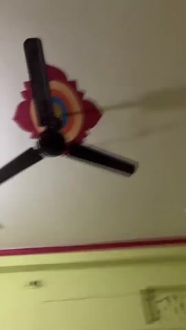 The video shows an indoor scene. The camera is moving and the view is blurry. A white ceiling fan with black blades is visible, along with what appears to be an air conditioning unit mounted high on a light green wall. There is also a patterned curtain or drapes visible. The room has a red and pink border along the top of the walls.
