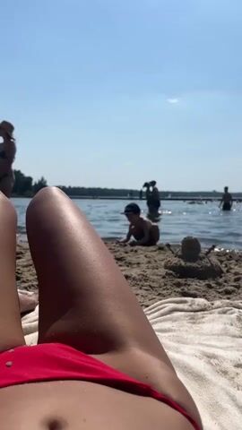 The video is a first-person perspective of someone lying on a sandy beach, wearing a red bikini. The camera is positioned low, focusing on the person's midsection and legs. In the background, a body of water, likely a lake or ocean, is visible. Several people are in the water, including children playing and adults. Some people are wading, while others appear to be swimming or relaxing. The sky is clear and blue, indicating a sunny day. The overall scene depicts a typical summer day at the beach with recreational activities.