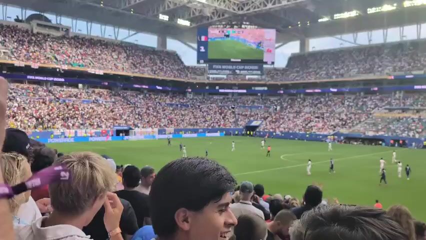 The video shows a soccer match in a large, packed stadium. The camera is positioned in the stands, looking down at the field. The scoreboard displays "FIFA WORLD CUP 2026". Players in white and blue uniforms are scattered across the green field. Spectators in the stands are wearing various colored clothing, some with their arms raised. The stadium has a modern roof structure. In the foreground, the heads and shoulders of several spectators are visible. The overall atmosphere appears to be one of excitement and engagement with the game.