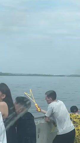 This is a vertical, medium-shot video of a group of people standing on what appears to be a boat or pier, looking out at a large body of water under an overcast sky. In the foreground, on the left, a person with long dark hair is partially visible, wearing a black top. Next to them, a person with dark hair is facing away from the camera, wearing a black hoodie. Between these two individuals, a person in a white t-shirt is holding a yellow bubble wand and blowing bubbles. The bubbles are visible in the air, some appearing to be in focus. On the far right, another person is partially visible, wearing a yellow shirt with a black pattern. The water is calm, with a distant shoreline visible on the horizon. The overall atmosphere is casual and relaxed.