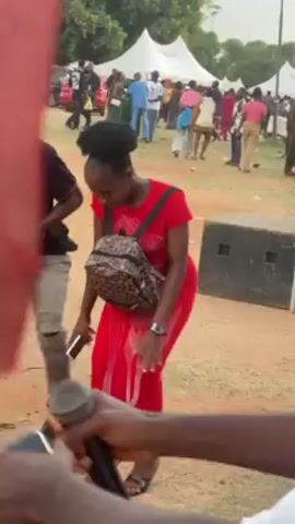 The video captures a lively outdoor event. A woman in a red dress with a leopard-print backpack is dancing in the center, surrounded by other people. The ground appears to be dirt or sand. Several tents are visible in the background, indicating a larger gathering. One man, wearing a colorful shirt and red shorts, dances near her, while others look on. The footage is somewhat shaky, suggesting it's taken with a handheld device, and someone is holding a red cloth with a name on it in front of the camera.