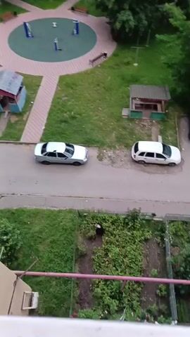 The video captures a bird in flight, its wings outstretched as it navigates the airspace above a residential area. Below, a small garden patch is visible, with a person tending to the plants. In the background, several cars are parked along a road that runs through the neighborhood. A playground with colorful equipment can be seen in the upper left corner, suggesting a family-friendly environment. The scene appears to be set during the daytime, with ample natural light illuminating the surroundings. The presence of greenery and the overall layout of the area point towards a suburban or urban setting in Karachayevsk, Russia. The mood is calm and observational, focusing on the natural element of the bird against the backdrop of human activity and infrastructure.
