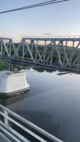 The video offers a picturesque view from a moving train, showcasing a metal truss bridge spanning a river. The scene is set in Zaluzhnoye, Russia, during what appears to be either sunrise or sunset, given the soft, warm light in the sky and the reflections on the water.

The camera initially captures a partial view through a train window, with a large, light-colored structure (possibly part of the train's interior or a door frame) temporarily obscuring much of the external landscape. As the train moves, this obstruction recedes, revealing a wide panorama.

The central subject is a grey metal truss bridge, characterized by its triangular and diagonal structural elements. Below the bridge, the river's surface is dark and remarkably reflective, mirroring the entire bridge structure and the soft, colorful hues of the sky. Green trees and foliage are visible along the riverbanks, adding to the natural beauty of the landscape. Thin overhead wires are also noticeable against the clear sky.

A voice is heard saying "Красиво" (Krasivo), which is Russian for "Beautiful," indicating admiration for the view. The overall impression is one of serene travel through a scenic environment during the golden hour.