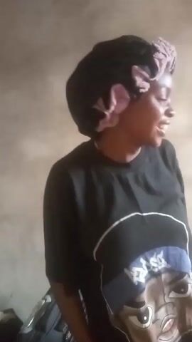 A woman wearing a black t-shirt and a pink and black hair bonnet is standing indoors against a neutral wall. She is looking off to the side and laughing.