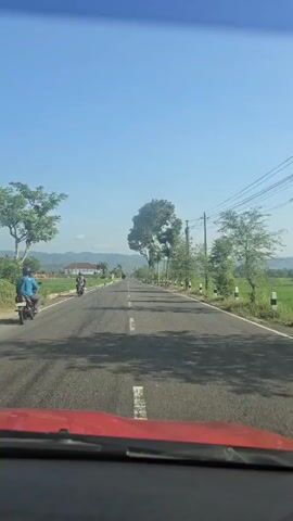A red car drives down a straight, paved road. There are two motorcycles ahead on the road. Trees line the road on the right. On either side of the road are fields of green vegetation. In the distance, there are mountains. The sky is blue and clear. There are utility poles and wires on the right side of the road.