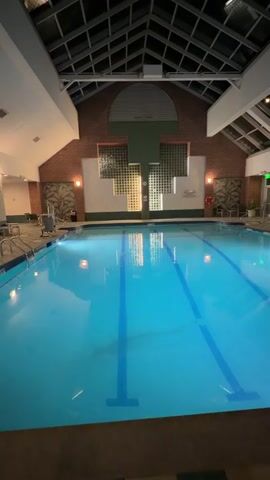 The video shows an indoor swimming pool with lanes. The pool is illuminated, and reflections of lights are visible on the water's surface. The architecture of the building includes a vaulted ceiling with a skylight and brick walls. There are some chairs and tables visible around the pool. The video appears to be a continuous shot of the pool area.