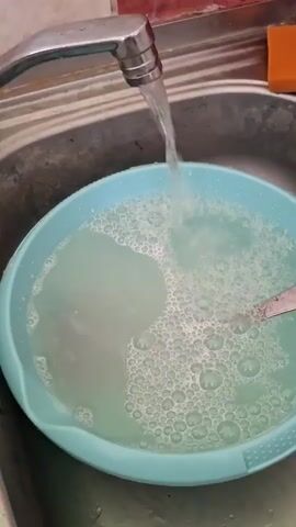 Water is running from a faucet into a light blue basin filled with soapy water. Bubbles are forming on the surface of the water. A spoon is visible in the basin. The sink basin is made of a grey, textured material. An orange sponge is visible on the edge of the sink.