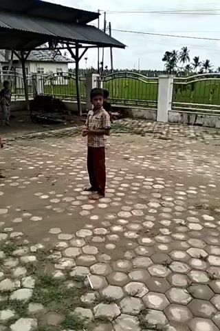 The video shows a group of children playing outside in a paved courtyard area. A child in a plaid shirt and maroon pants stands in the foreground, watching the others. Behind him, to the left, is a structure with a black roof. The other children are playing a game with a coconut. One child is shirtless. The playing area is paved with large, gray, hexagon-shaped stones. A white wall with a black iron fence runs along the back of the playing area, separating it from a field with palm trees in the distance. The sky is overcast.