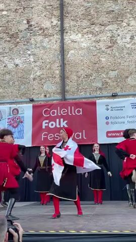 The video shows a folk dance performance at the Calella Folk Festival in Calella, Spain. Several young people in traditional costumes are dancing on a stage. The dancers on the left and right wear red and black outfits with gold trim, while the dancers in the center wear black dresses with red trim and headscarves. One male dancer in the center, wearing a red tunic and black pants, is prominently featured holding and twirling a flag with a red cross on a white background, which appears to be the flag of Georgia. The dancers are energetic and seem to be enjoying the performance.

In the background, a large banner reads "Calella Folk Festival." Above and to the right of this banner, there is text that appears to be from the "Ajuntament de Calella" (City Council of Calella) and mentions "Folklore Festes Calella" with social media icons and a website address "folklorefestescalella.com". The backdrop is a rough stone wall, suggesting an outdoor performance venue. The lighting appears to be daylight, likely afternoon, given the brightness and lack of artificial stage lighting. The overall atmosphere is festive and cultural.