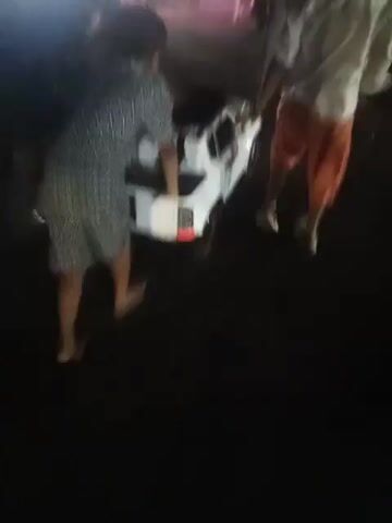 The video depicts a group of people moving a small, white, toy-sized pickup truck at night. Three people are directly involved in moving the truck, while others are nearby. One person is wearing a short-sleeved, patterned shirt, another is wearing a white shirt and orange capri pants, and a child in a blue shirt also helps move the truck. The scene is dimly lit, and the ground appears to be dark asphalt. There are some chairs and other objects visible in the background. The people are pushing and guiding the truck, presumably from one location to another.