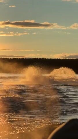 A person in a black wetsuit and knit cap stands on a bodyboard amidst splashing water, looking ahead towards a setting sun. In the background, another person rides a jet ski, kicking up spray, on a lake framed by a dark forest line. The scene is bathed in golden sunlight, with scattered clouds in the sky. The camera appears to be on another watercraft, moving through choppy water.