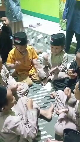 A group of young boys are sitting on the floor in a circle, some facing the camera and others facing away. They are all wearing traditional Islamic attire, including peci hats. The boys appear to be playing a game, with some clapping their hands and others watching intently. The environment appears to be indoors, possibly a home or a community gathering space, with a tiled floor and a green wall visible in the background. The lighting is dim, suggesting it might be evening or nighttime. The overall mood seems to be one of casual interaction and amusement among the children.