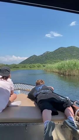 This video captures a serene moment aboard a boat navigating a narrow waterway surrounded by lush greenery and majestic mountains. The scene is bathed in bright daylight, indicating it's likely daytime, with a clear blue sky dotted with a few wispy clouds, suggesting fair weather.

The primary subjects are two individuals on the boat. In the foreground, a person wearing a pink t-shirt and a dark bucket hat is seated, facing away from the camera. To their right, another individual is lying face down on the boat's deck, also facing away, dressed in a dark t-shirt with a graphic design and black shorts. Their relaxed posture and the overall calm atmosphere suggest a leisurely experience.

The setting is a tranquil body of water, possibly a lake or a slow-moving river, bordered by dense reeds and vegetation on the right side of the frame. In the distance, rolling green mountains rise, their slopes covered in trees. The boat itself appears to be a modern, motorized vessel, with metallic accents and seating.

The activity is clearly a boat excursion, moving gently through the water, allowing the passengers to soak in the natural beauty of the landscape. The visual cues, such as the clear water, abundant vegetation, and mountainous backdrop, align with the provided location context of Prevlaka, Montenegro, known for its stunning natural scenery and waterways. There is no discernible text visible within the frame. The overall mood is peaceful and contemplative, a harmonious blend of human presence and the grandeur of nature.