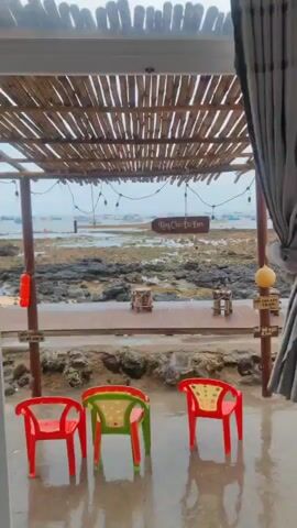 The video opens with a static shot of an outdoor sheltered area overlooking a rocky coastline. Three brightly colored plastic chairs (two red, one green) are in the foreground on wet pavement, suggesting recent rain. Above them, a bamboo-slatted roof is supported by wooden poles, from which string lights with bare bulbs are strung.

As the camera slowly pans right, the view reveals a rocky shore with puddles and shallow water, indicating either low tide or a very shallow bay. In the distance, numerous boats are visible on the water under an overcast sky. A wooden sign reads "Làng Chài Đá Đen," which translates to "Black Rock Fishing Village." Another sign, partially visible, seems to say "NƠI CÂY CỦA" (Place of the trees/roots). A third sign on a wooden pole reads "ĐẦU TRƯỜNG LÀ HÀ 500 M" (The arena is 500m).

At the 0:05 mark, a man is seen squatting on the rocky shore below the sheltered area, appearing to be engaged with something in his hands, possibly a phone. An orange tarp or cloth is also visible near him. An orange buoy hangs from one of the wooden support poles. The scene depicts a calm, overcast day in a coastal setting, likely in Phú Quý Special Administrative Region, Vietnam.