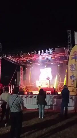 The video captures an outdoor nighttime event, likely a festival or concert. In the foreground, several people are seen from behind, facing a stage set up in the middle ground. The stage is illuminated with bright, colorful lights, including yellow and pink, and features a DJ booth with a DJ. A large screen behind the DJ displays a glowing graphic. Smoke or mist is visible around the stage, adding to the atmosphere. Beyond the stage, more people are gathered, and in the far background, various structures and tents are visible, along with more lighting. The overall setting appears to be an open, grassy area under a dark night sky.
