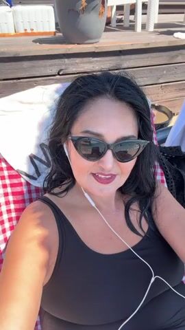 A woman with dark hair, sunglasses, and earbuds lies on a beach chair. She is wearing a black swimsuit and a red and white checkered blanket is draped over the chair. A colorful beach towel is also on the chair behind her. The scene appears to be a sunny day at the beach with wooden decking and some vegetation visible in the background.
