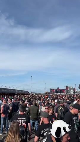 A handheld outdoor vertical shot captures a large crowd of people walking around at an outdoor festival during the daytime. The camera moves along with the crowd, offering a dynamic view of the festive atmosphere. Vendors' tents and stages are visible in the background, showing the various attractions of the event.