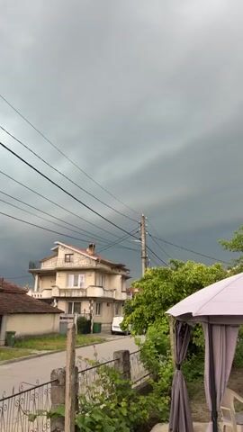 The video presents a static, eye-level view of a residential street in Dalgopol, Bulgaria, under a heavily overcast and dark sky, suggesting an approaching storm. In the foreground, a light-colored gazebo is visible on the right, partially covered by green foliage. To the left, a two-story house with a prominent balcony and orange-tiled roof stands along the street. A black car is visible on the street at the bottom left, partially obscured by a concrete fence, and appears to move slightly at the beginning of the clip. Further down the street, a white van is parked near the house. Numerous power lines stretch across the frame, connected to a utility pole positioned centrally. Green trees and other vegetation are scattered throughout the scene. No people are visible.