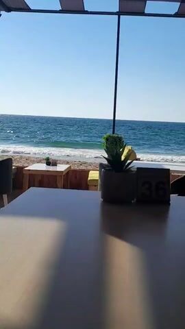 The video shows a view of the ocean from a table at an outdoor cafe. The camera is focused on the table in the foreground, with the beach and the ocean in the background. There are waves crashing on the shore, and the sky is clear and blue. A small potted plant is visible on the table, as well as a table number, which reads "36". The overall atmosphere appears to be relaxed and serene.