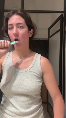 A woman in a light gray tank top stands facing the camera, brushing her teeth with a green and white toothbrush. She is indoors in what appears to be a bathroom. She has dark hair, fair skin, and a small turquoise pendant necklace. In the background, there is a dark-framed structure that could be a shower or a room divider. The scene is lit with an overhead light.