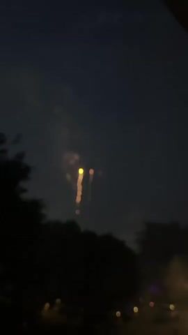 This video captures a nighttime scene with fireworks exploding in a dark sky above silhouetted trees and a street with lights. The fireworks are depicted as blurry, glowing orbs and streaks of light, with a variety of colors including yellow, orange, and white. The camera appears to be stationary, looking up at the sky. The overall atmosphere is festive and celebratory, consistent with a fireworks display. There are no people or discernible objects in the immediate foreground. The background consists of dark trees and some faint lights, suggesting a residential or suburban setting.