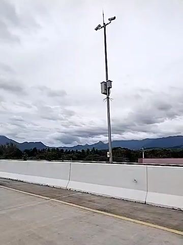 The video shows a highway with a white barrier alongside it. In the distance, there are mountains and a cloudy sky. It appears to be daytime, with the light diffused by the clouds. The road surface is concrete or asphalt. There are trees and other vegetation visible between the highway and the mountains. The video seems to be taken from a moving vehicle. A buzzing sound is heard.