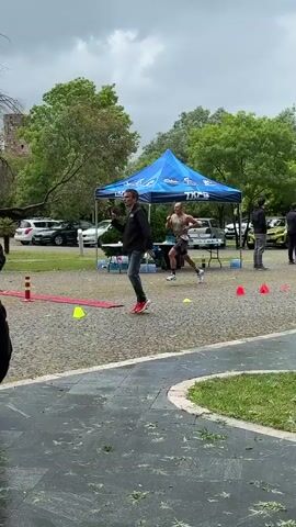 The video shows a man running through an obstacle course in a park. The course is marked with colored cones. The man is wearing running shorts and a tank top. He is running on a paved path. There are trees, cars, and people standing around the course. A blue tent is visible in the background. The weather appears cloudy and overcast. Some puddles are on the pavement.