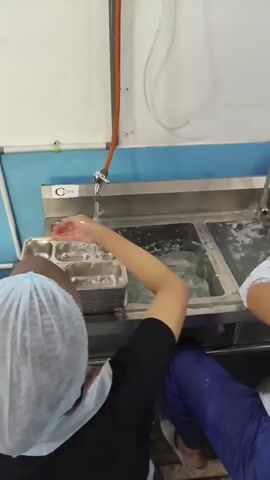 The video shows a group of people in what appears to be a commercial kitchen or food service area, engaged in washing dishes and trays. Multiple stainless steel sinks are visible, with water running and soap suds indicating cleaning activity. Several individuals are wearing hairnets and aprons, suggesting adherence to hygiene standards. In the foreground, stacks of stainless steel food trays are organized, some in large quantities. The environment seems functional and industrial, with stainless steel surfaces predominating. The actions depicted are consistent with a dishwashing or food preparation process.