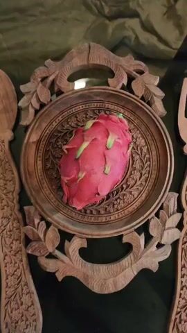 The video shows a close-up shot of a pink dragon fruit placed on a circular wooden plate with intricate carvings. The plate itself is ornate, with floral patterns and a decorative border. The dragon fruit has green and yellow tips on its scales. The background is a dark green fabric. The camera slowly zooms in and out on the fruit, highlighting its texture and color.