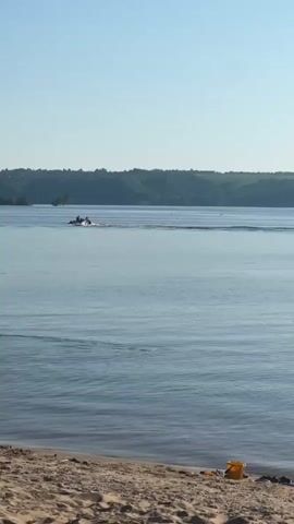The video shows a view of a wide river or lake on a clear, sunny day. The foreground features a sandy beach with a small yellow bucket. In the middle distance, there are three personal watercraft (jet skis) moving across the water. Each jet ski appears to have one or two people on board. Behind the watercraft, across the water, there is a shoreline covered with trees and vegetation, forming a continuous green hill. The sky is clear and blue, without any clouds. The water is mostly calm with some ripples and wakes created by the jet skis.