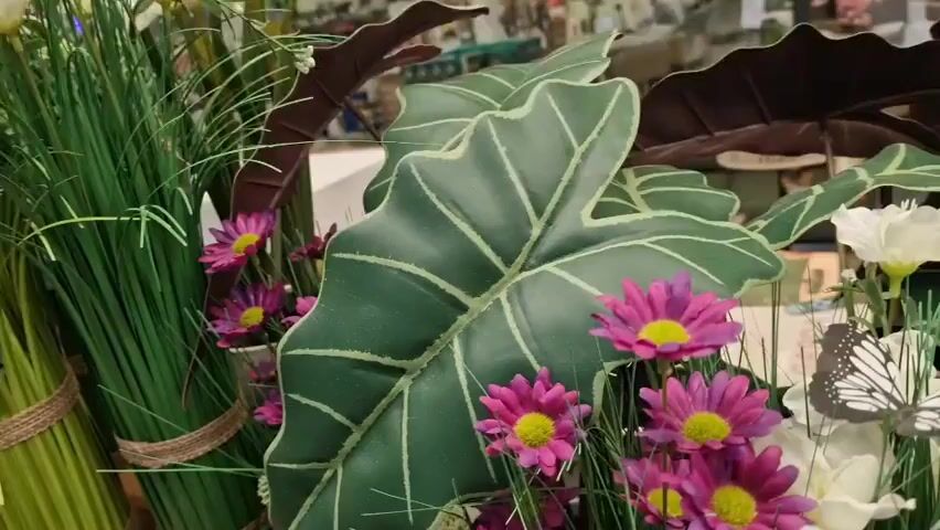 This is a close-up video of a floral arrangement. The arrangement features a variety of artificial flowers, including pink daisies with yellow centers, white flowers, and pink tulips. There are also large, dark green leaves with prominent white veins, and tall, green grass-like foliage. Several artificial butterflies with black and white patterns are perched on the flowers and foliage. The background is blurred, but appears to be an indoor setting, possibly a store or home, with shelves and other decorative items visible. The lighting is bright and even, illuminating the details of the artificial plants and flowers.