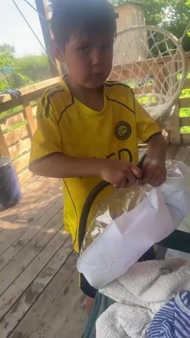 A young boy in a yellow soccer jersey with black accents is standing on a wooden deck, holding a black hose and a clear plastic object. The boy is looking at the object with a focused expression. The object appears to be an inflatable, possibly a ring or a float, with white material inside. The boy's hands are engaged with the hose and the object, suggesting he is inflating it.

The setting is outdoors, likely a backyard or a porch, with wooden planks forming the floor of the deck. In the background, a white netted hanging chair is visible, along with a wooden fence and lush green foliage. The lighting suggests it is daytime, with sunlight casting shadows. The overall mood is casual and active, with the boy engaged in a task.

Visible text on the boy's jersey includes the word "KAED" and a circular logo with some emblem. The hose he is holding is black and appears to be connected to an inflation device, although the device itself is not fully visible. The inflatable object has a clear outer layer and a white inner part, with a textured or patterned surface on the inner white material. A fluffy white and blue striped towel is visible in the lower right corner of the frame, resting on a green surface.