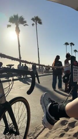 The video opens with a close-up on a person's leg and foot, wearing a white sneaker and black socks, resting on a sandy surface. A bicycle is visible in the foreground on the left, with its handlebars and front wheel in focus. The background reveals a sunny beach scene with palm trees swaying under a clear blue sky. In the mid-ground, people are walking along a boardwalk, and a small, three-wheeled vehicle passes by. A rope barrier runs horizontally across the frame, separating the sandy area from the boardwalk. The sun is visible in the upper left corner, casting a bright light. The scene appears to be a lively outdoor setting, likely a beach town, during daytime.