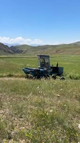 A blue, agricultural machine, possibly a harvester, is positioned in a grassy field under a clear blue sky with scattered white clouds. The machine is stationary and appears to be facing slightly to the left of the camera. In the background, rolling green hills form a natural landscape, with some visible geological formations and what might be power lines on the horizon. The field is covered in green grass and small yellow and white wildflowers. The machine has a cab for the operator and a large harvesting apparatus at the front. The overall scene depicts a rural, possibly agricultural, setting on a sunny day.