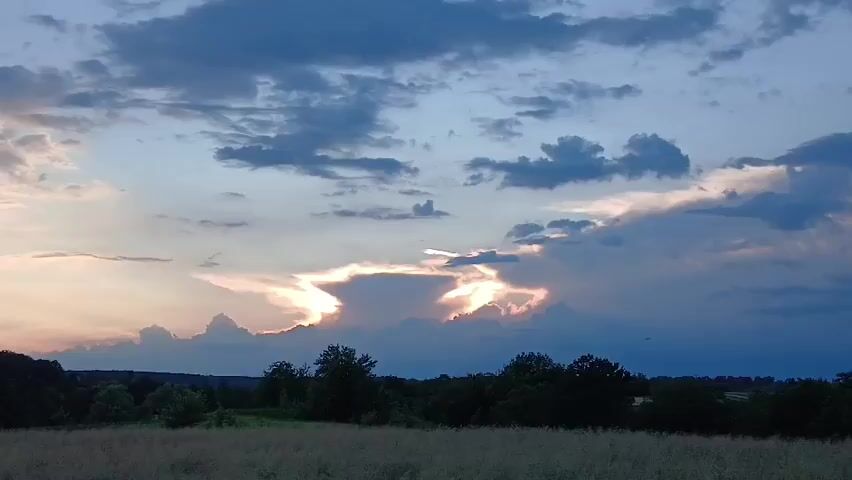 A time lapse video captures the changing sky at dusk. Dark clouds drift across a pale blue sky, with the sun creating a bright, glowing aperture between them. A line of trees forms the horizon, silhouetted against the colorful sky, with a field of tall, dry grass in the foreground.