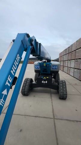The video shows a Genie Z33/18 boom lift on a concrete surface in what appears to be a commercial yard. The lift is blue and gray, with "TE HUUR" written on the side of the boom, which means "FOR RENT" in Dutch. It has a 4x4 drive and is equipped with tracks instead of wheels. In the background, there are stacks of wooden crates or pallets. The sky is overcast. The boom of the lift appears to be moving up slightly.