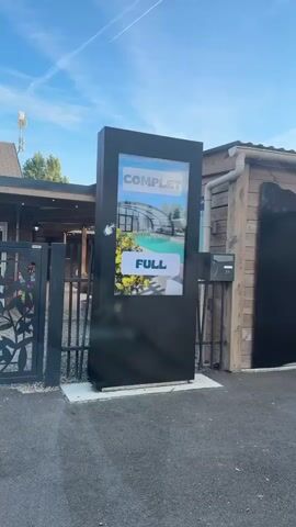 The video shows an outdoor digital display screen placed at the entrance of a facility, likely a campground or resort. The screen displays the words "COMPLET" and "FULL" in large white lettering against a background image of a covered swimming pool area. To the right of the screen is a wooden building with the words "Camping Star" and some stars visible on its side. There is also a mailbox attached to the building. In the background, there is a gate with a decorative leaf pattern. The sky is visible with some clouds and contrails from an airplane. The overall scene suggests that the facility is full and not accepting new visitors.