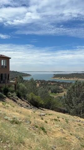 A vertical video shows a view of a large lake under a partly cloudy blue sky. The foreground is a dry, grassy hillside with a few scattered rocks. A fence runs along the edge of the slope. In the middle ground, there are numerous trees and some buildings partially visible. The lake extends into the background, with treed hills surrounding it. The water appears calm. The camera pans slowly to the right.
