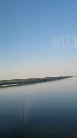 The video shows a wide, calm river under a clear blue sky. The camera appears to be moving, possibly from a vehicle like a train or boat, as evidenced by the blurry vertical lines reflected in the water, resembling window blinds or curtains. Along the far bank of the river, there is a line of trees and vegetation. The water is dark blue and reflects the sky and the distant treeline. The overall impression is one of peaceful travel through a natural landscape.
