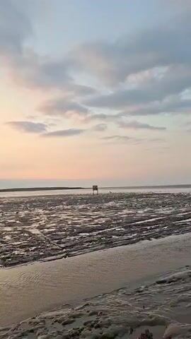The video shows a beach at sunset. The sky is filled with clouds, and the sun is setting over the water. The tide is out, revealing a muddy, textured shoreline. In the distance, a small wooden structure stands on stilts. Closer to the camera, there are remnants of a brick structure, possibly ruins. A person wearing a dark t-shirt, light-colored shorts, and a baseball cap stands with their arms crossed, looking out at the sea. In the background, other people and a motorcycle are visible. The video pans slightly to reveal more of the beach and the surrounding vegetation.
