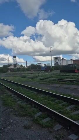 A video pans from left to right across a railway yard. The foreground consists of two sets of railway tracks, with gravel and grass along the sides. In the mid-ground, there are more tracks, grass, and some trees. Beyond the tracks, in the background, there are several industrial buildings, a water tower, and a blue and red train engine. A green train car is also visible on the right. The sky is blue with large white clouds.